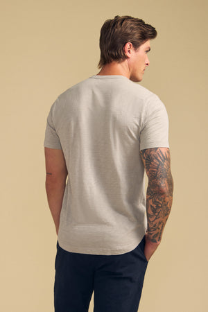 A man stands with his back to the camera, hands in pockets, revealing a tattoo on his right arm while wearing the AMARO TEE by Velvet by Graham & Spencer— a light gray Peruvian cotton slub crew-neck short-sleeve t-shirt— and dark pants against a beige background.