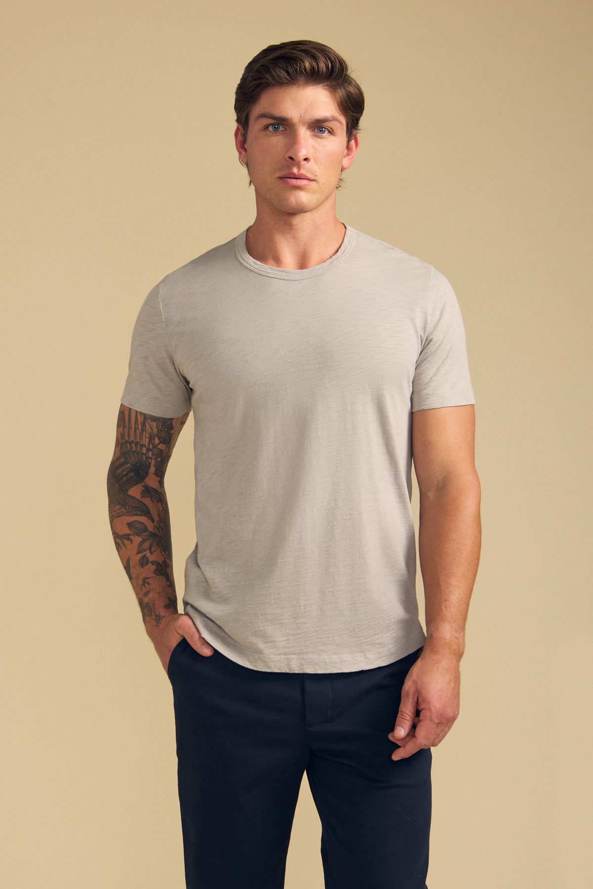 A man with short hair and a tattoo on his left arm wears the AMARO TEE by Velvet by Graham & Spencer, a light beige crew neck Peruvian cotton slub t-shirt, paired with dark pants, standing against a plain beige background.
