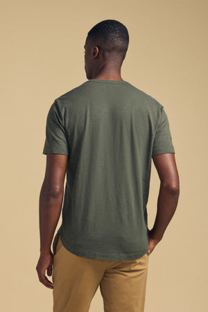 A man stands with his back to the camera, wearing the AMARO TEE by Velvet by Graham & Spencer—an olive green Peruvian cotton t-shirt featuring a classic crew neckline—paired with tan pants against a plain beige background. Ideal as a layering piece for versatile styling.