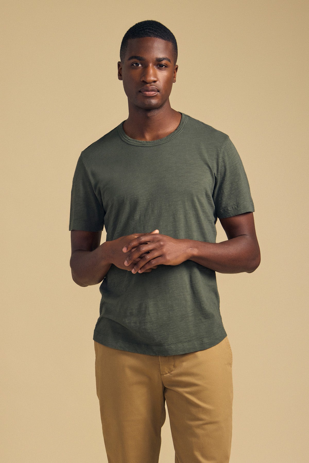 A man stands against a beige background, wearing Velvet by Graham & Spencer’s AMARO TEE—an olive green Peruvian cotton t-shirt featuring a classic crew neckline—paired with tan pants and his hands loosely clasped in front of him, making it a perfect layering piece for any season.