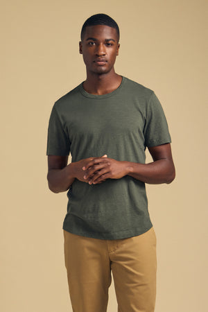 A man stands against a beige background, wearing Velvet by Graham & Spencer’s AMARO TEE—an olive green Peruvian cotton t-shirt featuring a classic crew neckline—paired with tan pants and his hands loosely clasped in front of him, making it a perfect layering piece for any season.