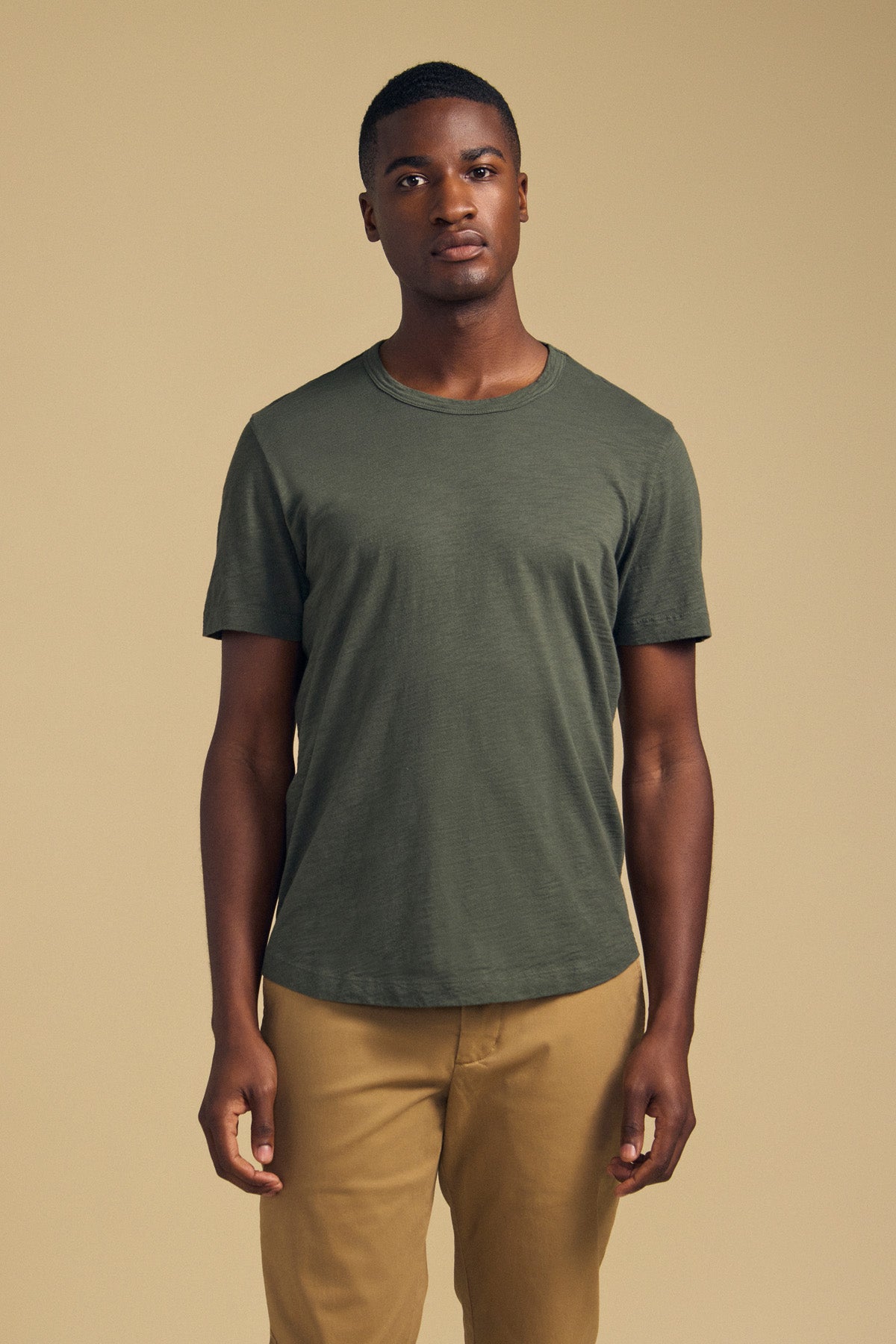 A person stands facing forward wearing the AMARO TEE by Velvet by Graham & Spencer—a short-sleeve t-shirt in olive green, crafted from soft Peruvian cotton—paired with tan pants against a plain beige background. Featuring a classic crew neckline, this versatile tee is an ideal layering piece for any wardrobe.