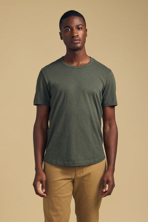 A person stands facing forward wearing the AMARO TEE by Velvet by Graham & Spencer—a short-sleeve t-shirt in olive green, crafted from soft Peruvian cotton—paired with tan pants against a plain beige background. Featuring a classic crew neckline, this versatile tee is an ideal layering piece for any wardrobe.