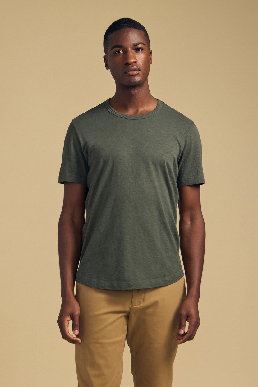A person stands facing forward wearing the AMARO TEE by Velvet by Graham & Spencer—a short-sleeve t-shirt in olive green, crafted from soft Peruvian cotton—paired with tan pants against a plain beige background. Featuring a classic crew neckline, this versatile tee is an ideal layering piece for any wardrobe.