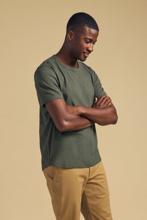 A man stands with arms crossed, looking down and smiling. He wears the AMARO TEE by Velvet by Graham & Spencer—a green crew neckline t-shirt crafted from soft Peruvian cotton—paired with tan pants against a plain beige background. This tee is a perfect layering piece for any casual outfit.