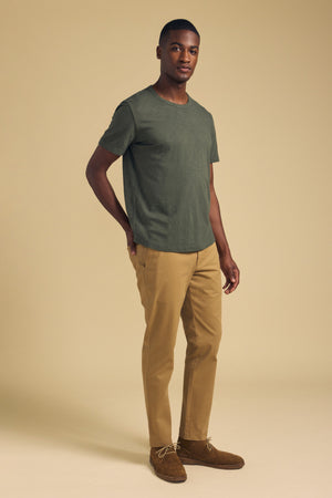 A man stands against a beige background wearing the Velvet by Graham & Spencer AMARO TEE in olive green Peruvian cotton with a crew neckline, paired with tan pants and brown suede shoes.