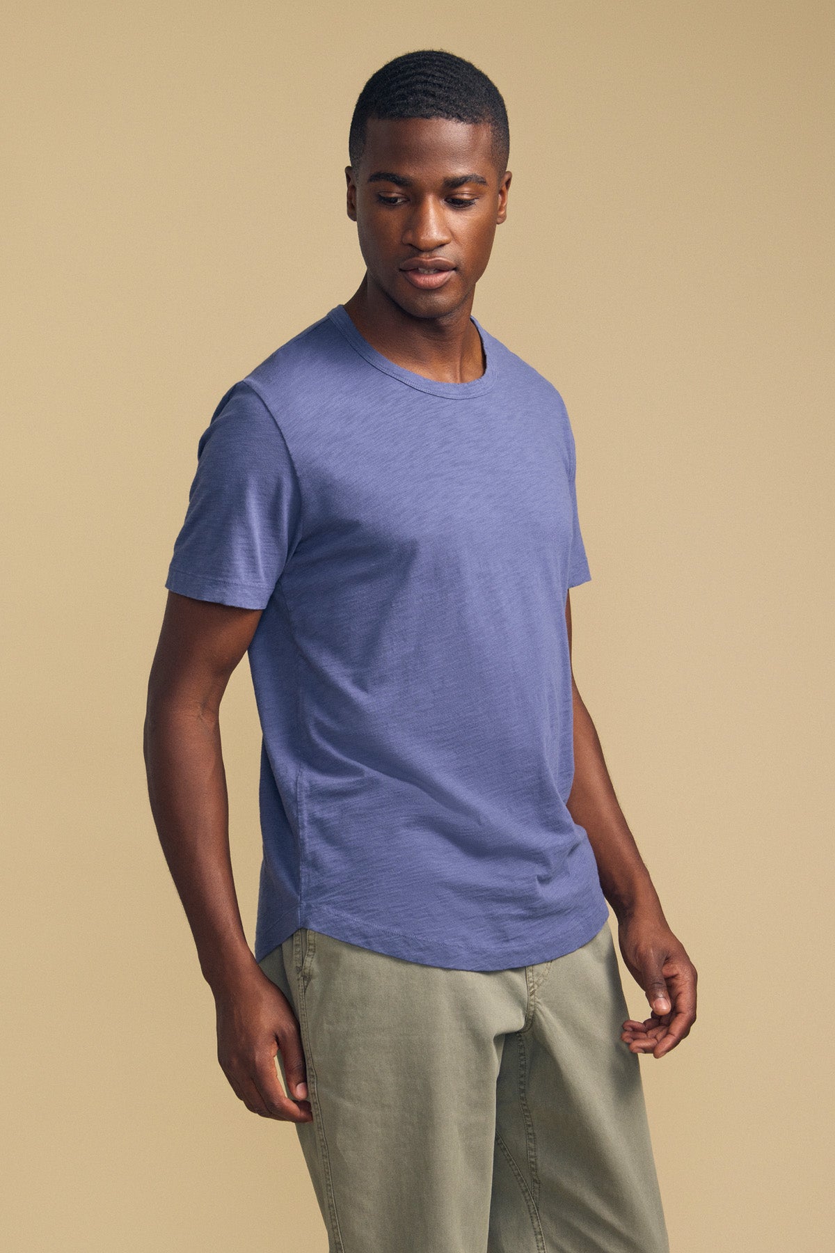 A man stands against a plain beige background, looking slightly to his left, dressed in the AMARO TEE by Velvet by Graham & Spencer—a blue short-sleeve t-shirt crafted from soft Peruvian cotton with a classic crew neckline—and light khaki pants. The versatile design of the AMARO TEE makes it an ideal layering piece for any casual look.