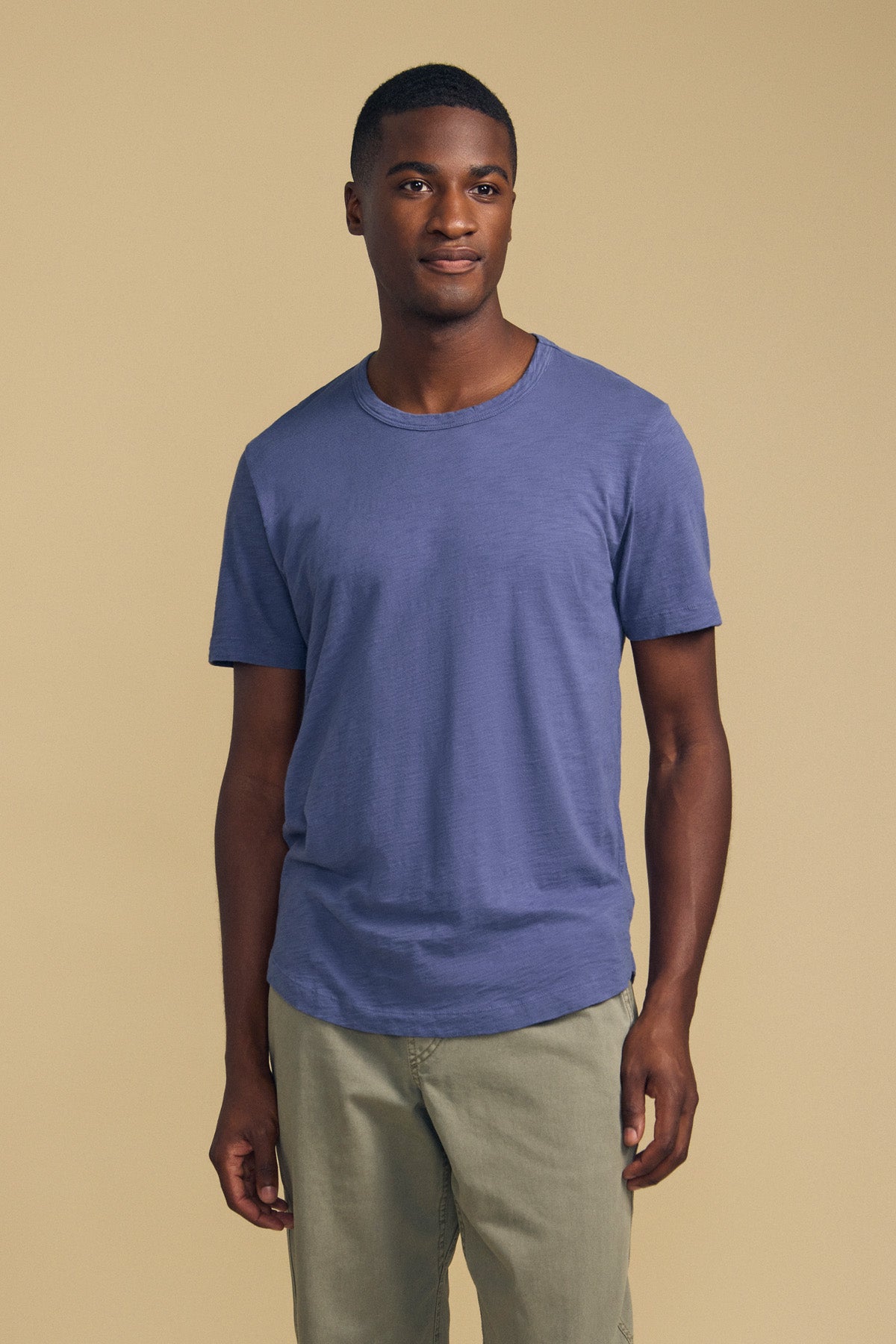 A person stands against a plain beige background, wearing the AMARO TEE by Velvet by Graham & Spencer—a short-sleeve blue t-shirt crafted from soft Peruvian cotton with a classic crew neckline—paired with light khaki pants, looking slightly to the side with a neutral expression.