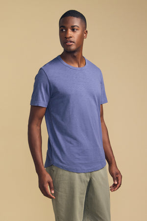 A man stands against a plain beige background, wearing the AMARO TEE by Velvet by Graham & Spencer—a blue short-sleeve t-shirt made from Peruvian cotton—paired with light green pants and looking slightly to his left. The crew neckline of the AMARO TEE makes it an ideal layering piece for effortless style.