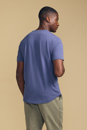 A man stands with his back to the camera, wearing the AMARO TEE from Velvet by Graham & Spencer—a short-sleeve blue t-shirt with a classic crew neckline—paired with light green pants against a beige background.