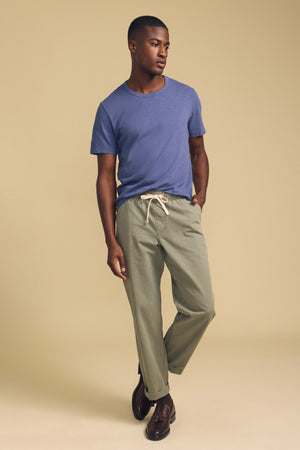 A man stands against a beige background, wearing the AMARO TEE by Velvet by Graham & Spencer—a blue Peruvian cotton T-shirt with a classic crew neckline—paired with light olive-green drawstring pants and dark brown shoes, with one hand in his pocket.
