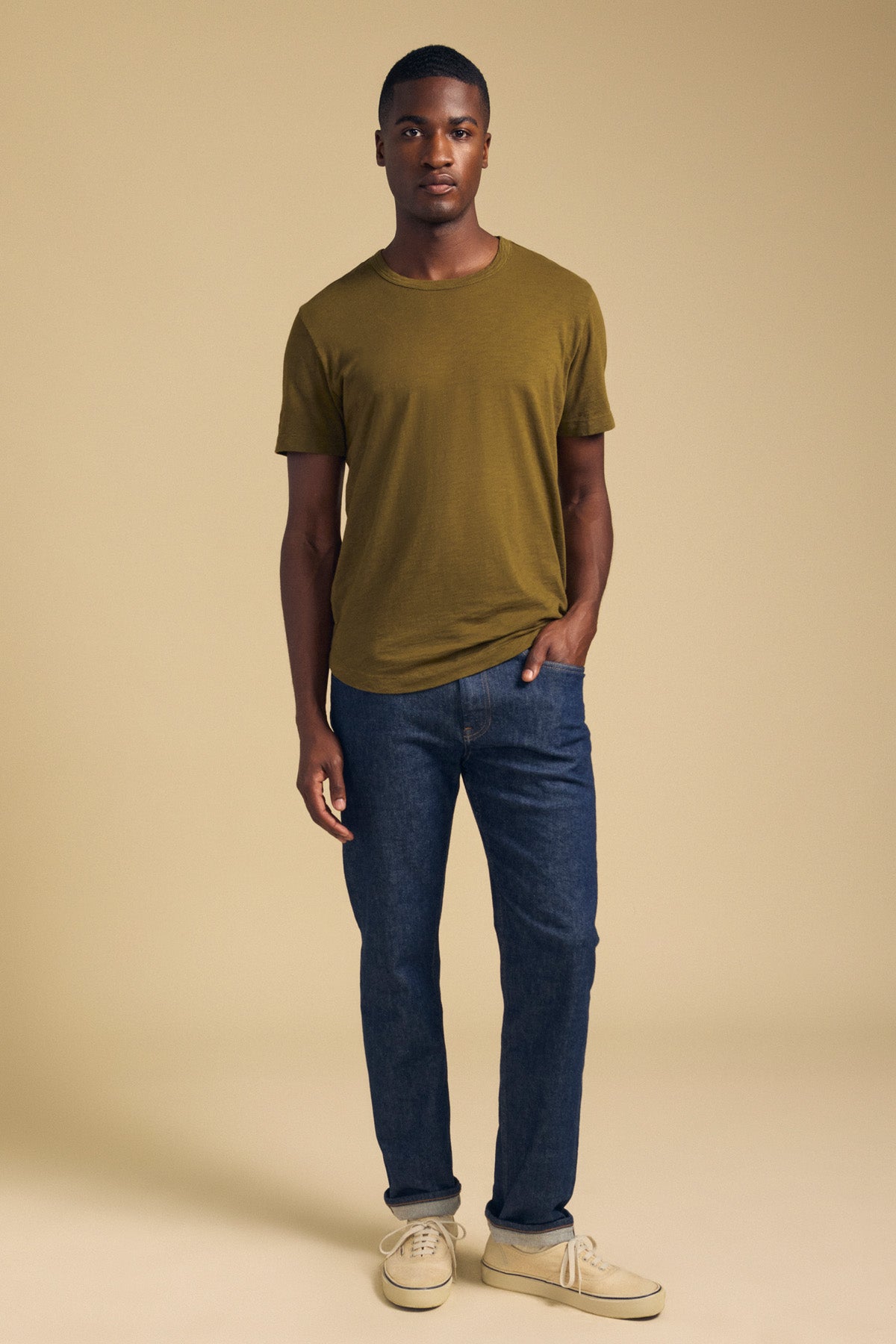 A man wearing an olive green AMARO TEE in Peruvian cotton with a crew neckline, blue jeans, and tan sneakers.