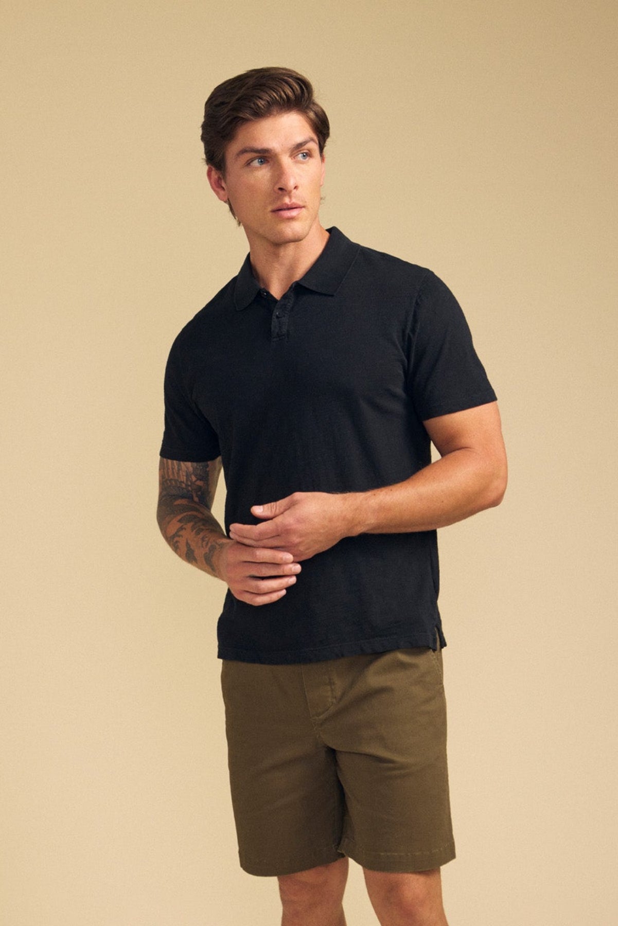 A man wears the MORRIS POLO by Velvet by Graham & Spencer, a black cotton slub polo with a ribbed collar and short sleeves, paired with olive green shorts, standing against a neutral beige background.