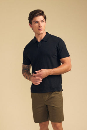 A man wears the MORRIS POLO by Velvet by Graham & Spencer, a black cotton slub polo with a ribbed collar and short sleeves, paired with olive green shorts, standing against a neutral beige background.