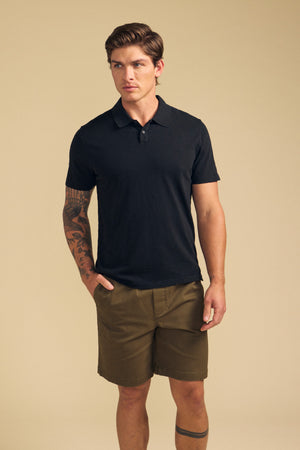 A man stands with hands in pockets, wearing the Velvet by Graham & Spencer MORRIS POLO in black and olive green shorts, displaying a right arm tattoo against a plain beige background.