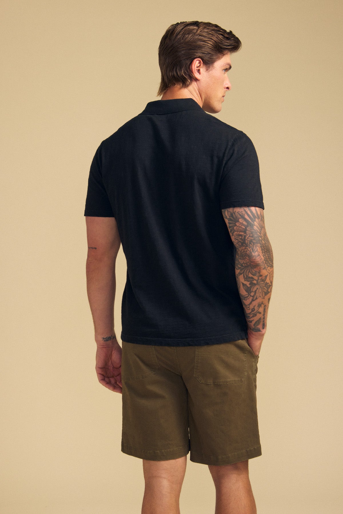A man faces away, wearing the Velvet by Graham & Spencer MORRIS POLO in black, paired with olive green shorts. His tattooed right arm is visible against a plain beige background.