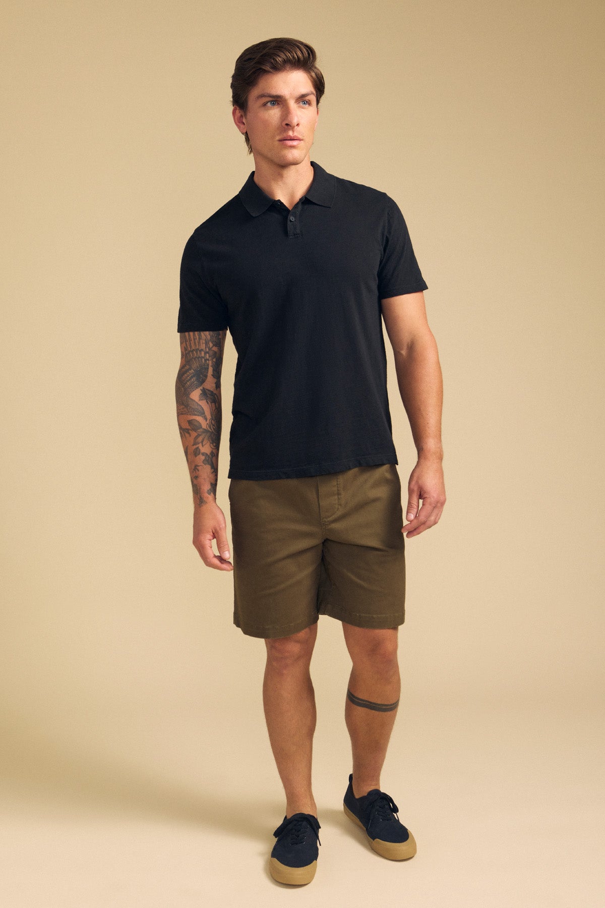 A man with tattoos on his left arm and lower right leg stands against a beige background, wearing the black MORRIS POLO by Velvet by Graham & Spencer, olive green shorts, and black sneakers.-41702106955969