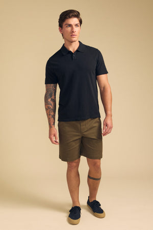 A man with tattoos on his left arm and lower right leg stands against a beige background, wearing the black MORRIS POLO by Velvet by Graham & Spencer, olive green shorts, and black sneakers.
