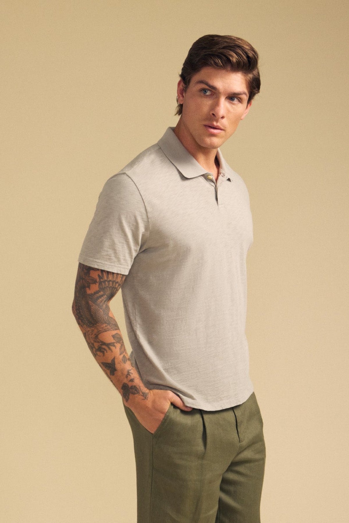 A man wearing the Velvet by Graham & Spencer MORRIS POLO in light gray cotton slub and olive green pants stands with one hand in his pocket against a beige background.