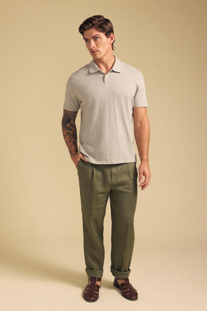 A man stands against a plain background, wearing the MORRIS POLO by Velvet by Graham & Spencer in light beige with a ribbed collar, paired with olive green trousers and brown sandals; he has one hand in his pocket and a tattoo on his right arm.