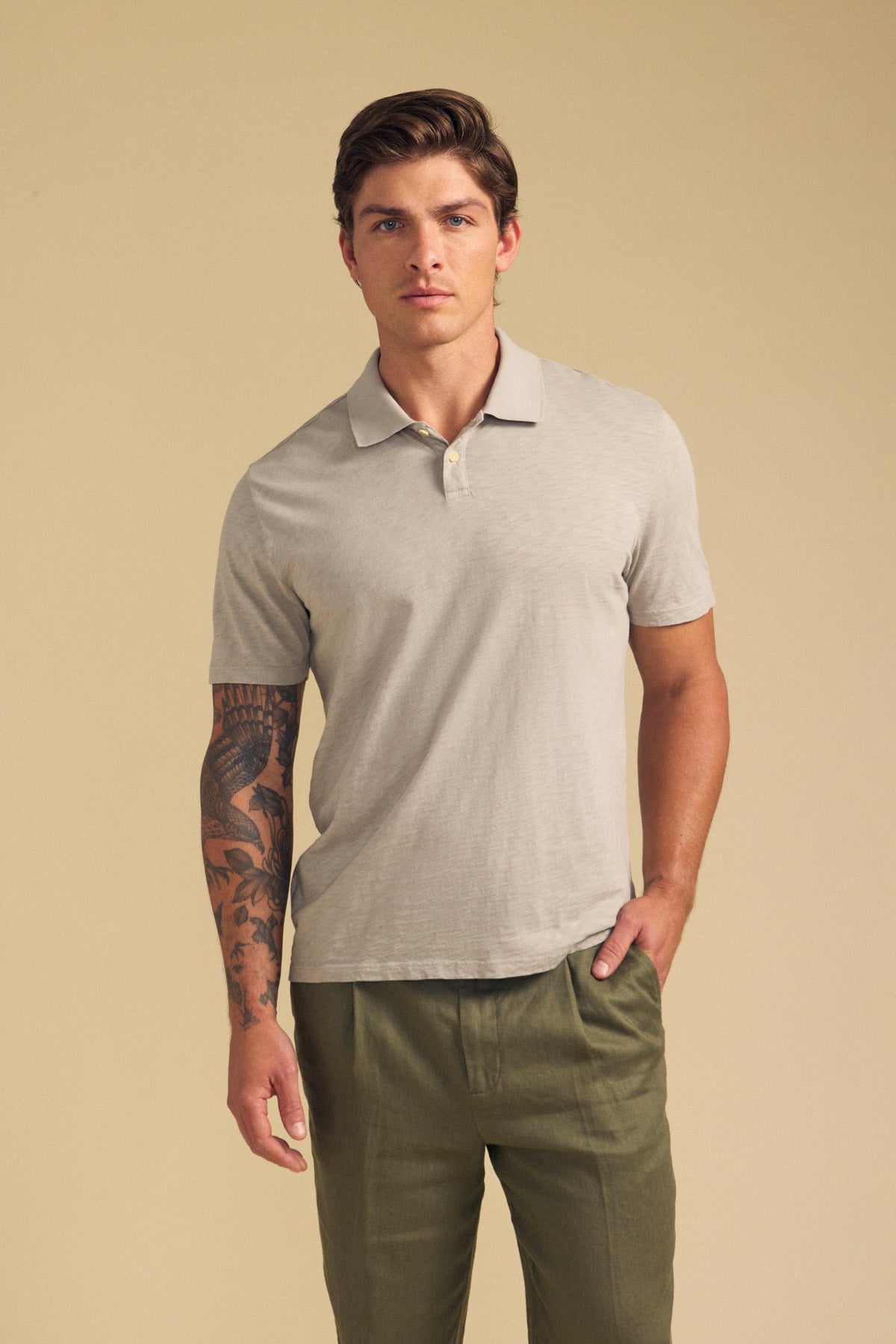 A man wearing the MORRIS POLO by Velvet by Graham & Spencer in light grey cotton slub with short sleeves and olive green pants stands against a beige background, showing a tattoo on his left arm.-41702106726593