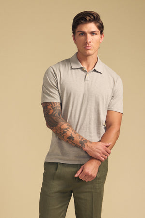 A man with short brown hair and a tattooed right arm wears the Velvet by Graham & Spencer MORRIS POLO in light grey and olive green pants, standing against a plain beige background.