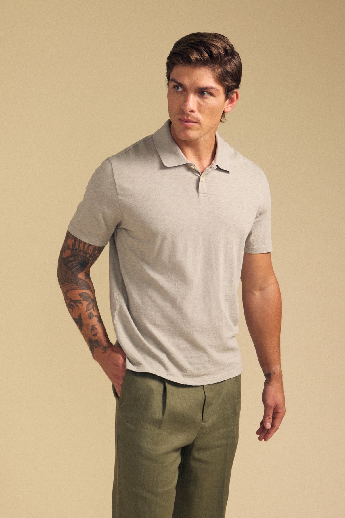 A man stands against a beige background, wearing the MORRIS POLO by Velvet by Graham & Spencer—a light beige cotton slub polo with short sleeves and ribbed collar—paired with olive green pants, one hand in his pocket.-41702106628289