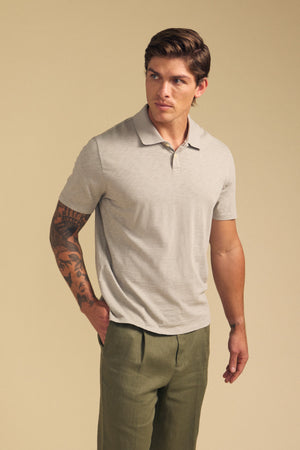 A man stands against a beige background, wearing the MORRIS POLO by Velvet by Graham & Spencer—a light beige cotton slub polo with short sleeves and ribbed collar—paired with olive green pants, one hand in his pocket.