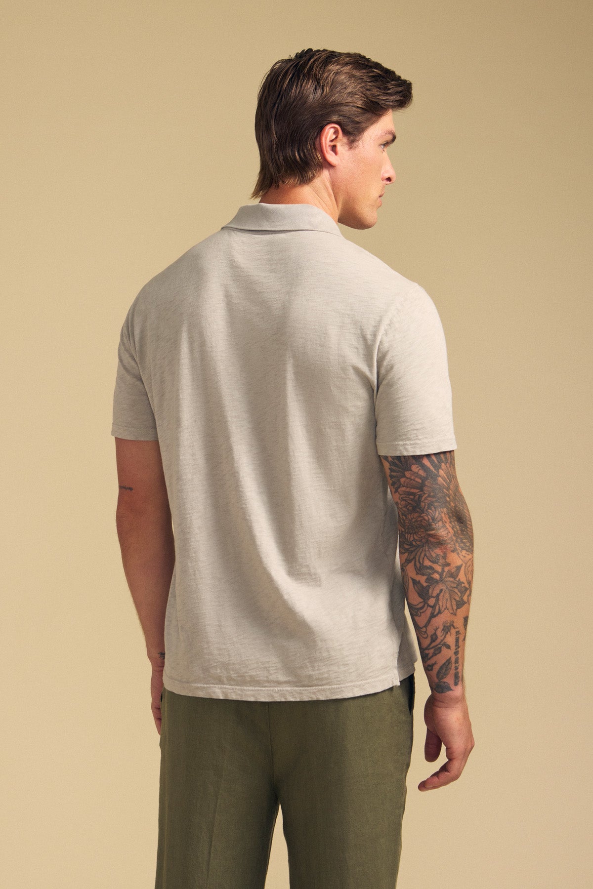 A man stands with his tattooed right arm visible, wearing the Velvet by Graham & Spencer MORRIS POLO in light gray cotton slub with short sleeves and a ribbed collar, paired with olive green pants, against a plain beige background.-41702106562753