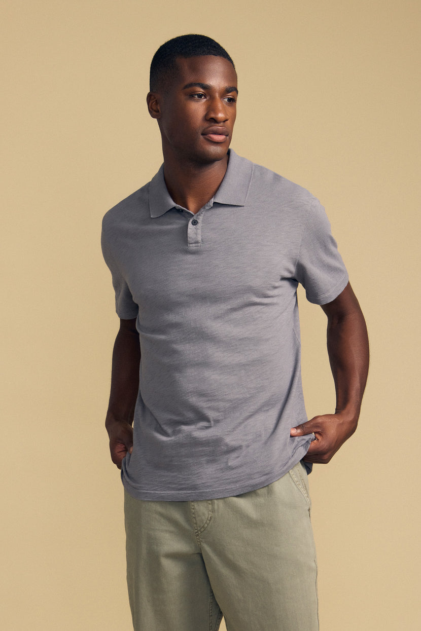 Wearing the MORRIS POLO by Velvet by Graham & Spencer—a light gray, short-sleeve cotton slub polo with a ribbed collar—paired with light khaki pants, a person stands against a plain beige background, gazing slightly to the side.
