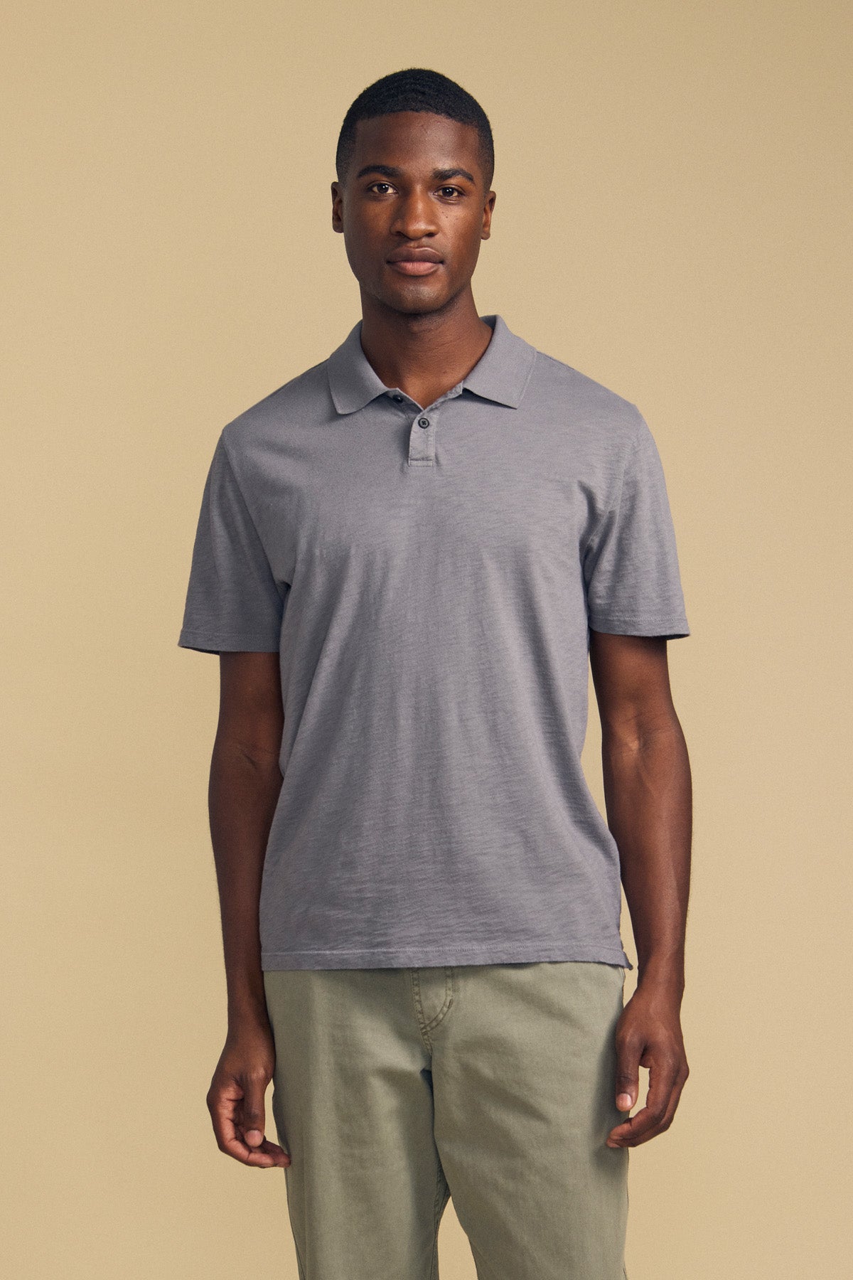 A man stands facing forward, wearing the MORRIS POLO by Velvet by Graham & Spencer—a short-sleeved gray cotton slub polo with a ribbed collar—paired with light green pants against a plain beige background.-41859748888769