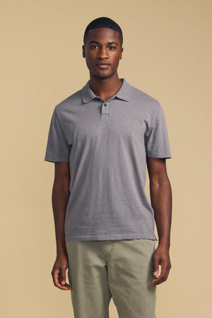 A man stands facing forward, wearing the MORRIS POLO by Velvet by Graham & Spencer—a short-sleeved gray cotton slub polo with a ribbed collar—paired with light green pants against a plain beige background.