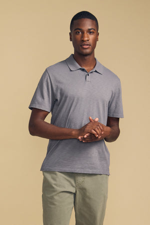 A person stands against a beige background, wearing the MORRIS POLO by Velvet by Graham & Spencer—a cotton slub polo with a ribbed collar and short sleeves—paired with light khaki pants, hands clasped in front.