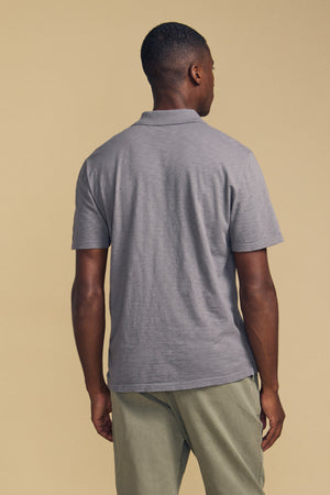 A man stands facing away, wearing the Velvet by Graham & Spencer MORRIS POLO—a short-sleeve cotton slub polo with a ribbed collar—paired with light green pants against a beige background.
