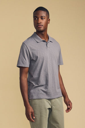 A man stands facing slightly to the side, wearing the MORRIS POLO by Velvet by Graham & Spencer—a grey cotton slub polo with a ribbed collar and short sleeves—paired with light green pants, against a plain beige background.