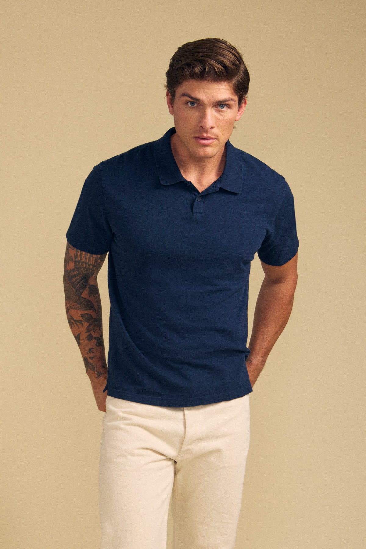 A man with short brown hair and a left arm tattoo wears the MORRIS POLO by Velvet by Graham & Spencer in navy, featuring a ribbed collar and short sleeves, paired with light beige pants, against a plain beige background.-41702104006849