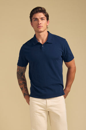 A man with light brown hair, wearing the MORRIS POLO by Velvet by Graham & Spencer in navy blue and beige pants, stands against a plain beige background. One arm features a visible tattoo.