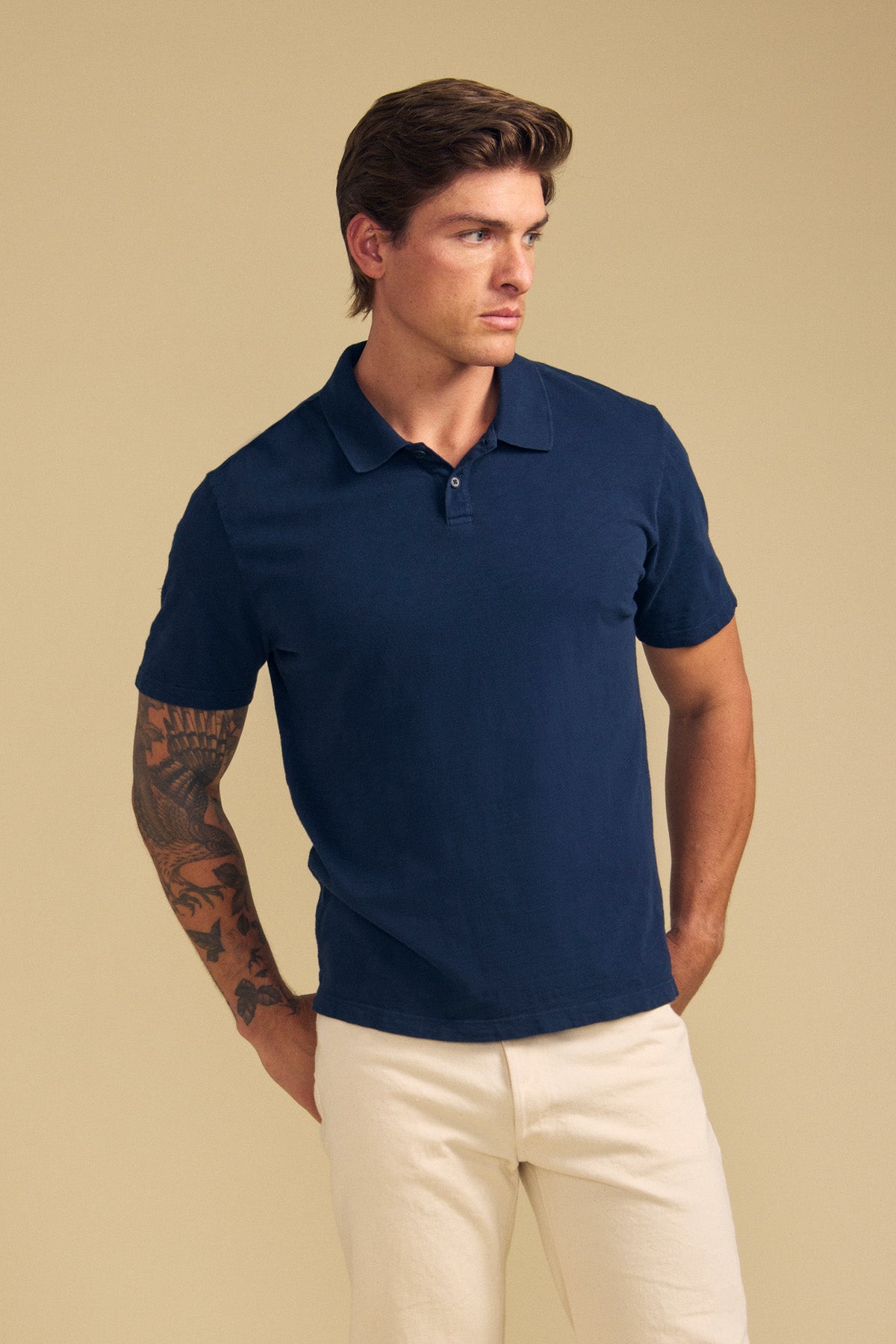 A man with a tattoo on his left arm wears the MORRIS POLO by Velvet by Graham & Spencer in navy blue with cream pants, standing against a plain beige background.