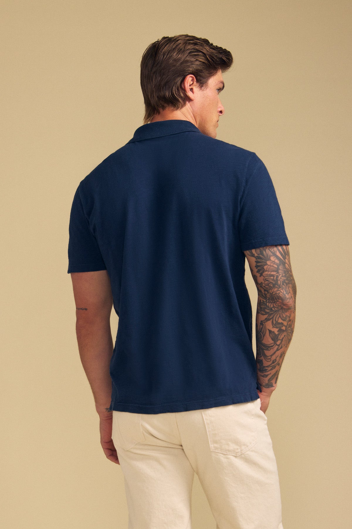 A person with short brown hair is seen from the back, wearing Velvet by Graham & Spencer's MORRIS POLO in navy blue with short sleeves and light-colored pants, set against a plain beige background.