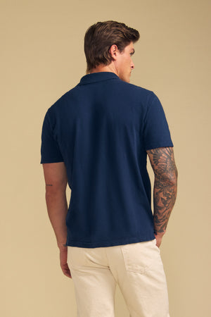 A person with short brown hair is seen from the back, wearing Velvet by Graham & Spencer's MORRIS POLO in navy blue with short sleeves and light-colored pants, set against a plain beige background.