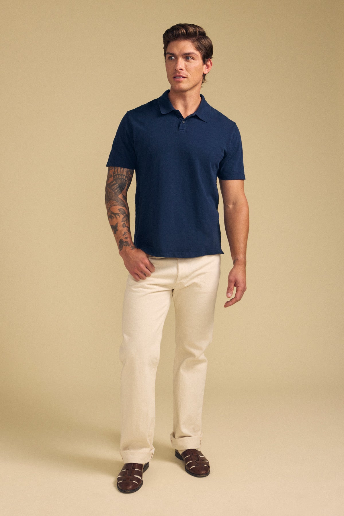 A man stands against a beige background, wearing the Velvet by Graham & Spencer MORRIS POLO in navy with a ribbed collar and two-button placket, paired with light beige pants and brown sandals. A tattoo is visible on one arm.