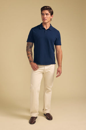 A man stands against a beige background, wearing the Velvet by Graham & Spencer MORRIS POLO in navy with a ribbed collar and two-button placket, paired with light beige pants and brown sandals. A tattoo is visible on one arm.