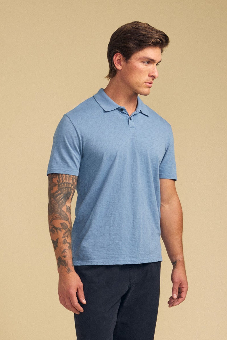 A man with short brown hair and a left forearm tattoo wears the MORRIS POLO by Velvet by Graham & Spencer—a light blue cotton slub polo with a ribbed collar—paired with dark pants, standing against a beige background.-41702102106305