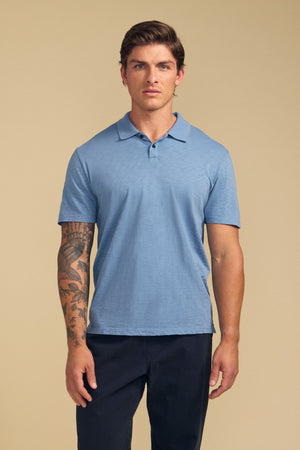 A man wears the MORRIS POLO by Velvet by Graham & Spencer—a light blue cotton slub, short-sleeved shirt—with dark pants, standing facing forward against a plain beige background.