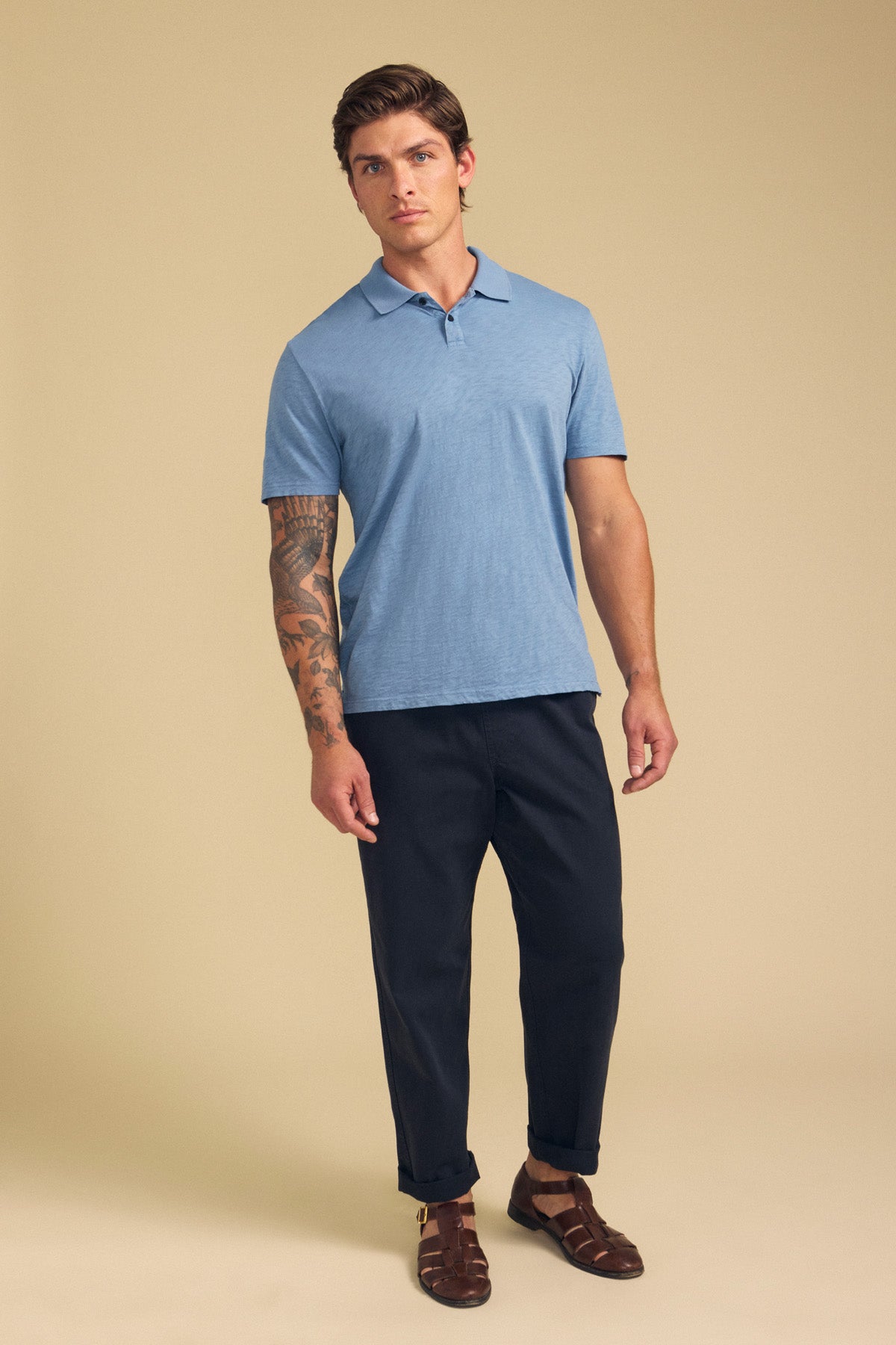 A man with a visible arm tattoo and neutral expression stands against a beige background wearing the MORRIS POLO by Velvet by Graham & Spencer, styled in light blue cotton slub with a ribbed collar, paired with dark pants and brown sandals.