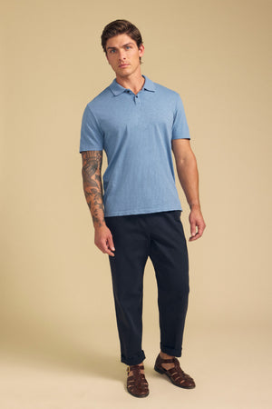 A man with a visible arm tattoo and neutral expression stands against a beige background wearing the MORRIS POLO by Velvet by Graham & Spencer, styled in light blue cotton slub with a ribbed collar, paired with dark pants and brown sandals.
