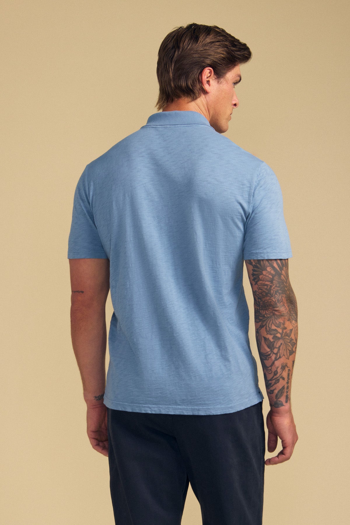 A man stands with his back to the camera, wearing the Velvet by Graham & Spencer MORRIS POLO in light blue with short sleeves and a ribbed collar, paired with dark pants and showing a tattoo sleeve on his left arm against a beige background.-41702101909697