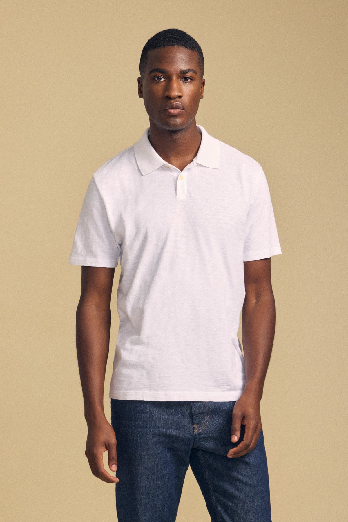 A person wearing the MORRIS POLO by Velvet by Graham & Spencer—a white cotton slub polo with a ribbed collar and short sleeves—paired with blue jeans, stands against a plain beige background facing the camera neutrally.