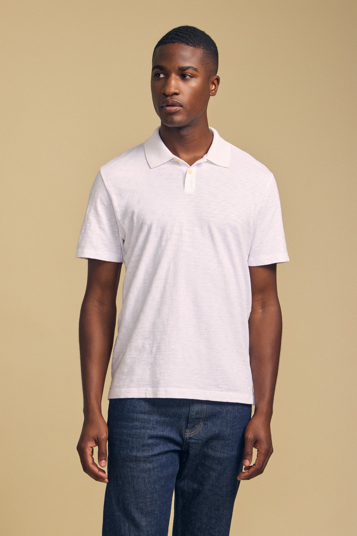A man faces forward wearing the MORRIS POLO by Velvet by Graham & Spencer— a plain white short-sleeve cotton slub polo with a ribbed collar—paired with dark jeans against a beige background.-41702098600129
