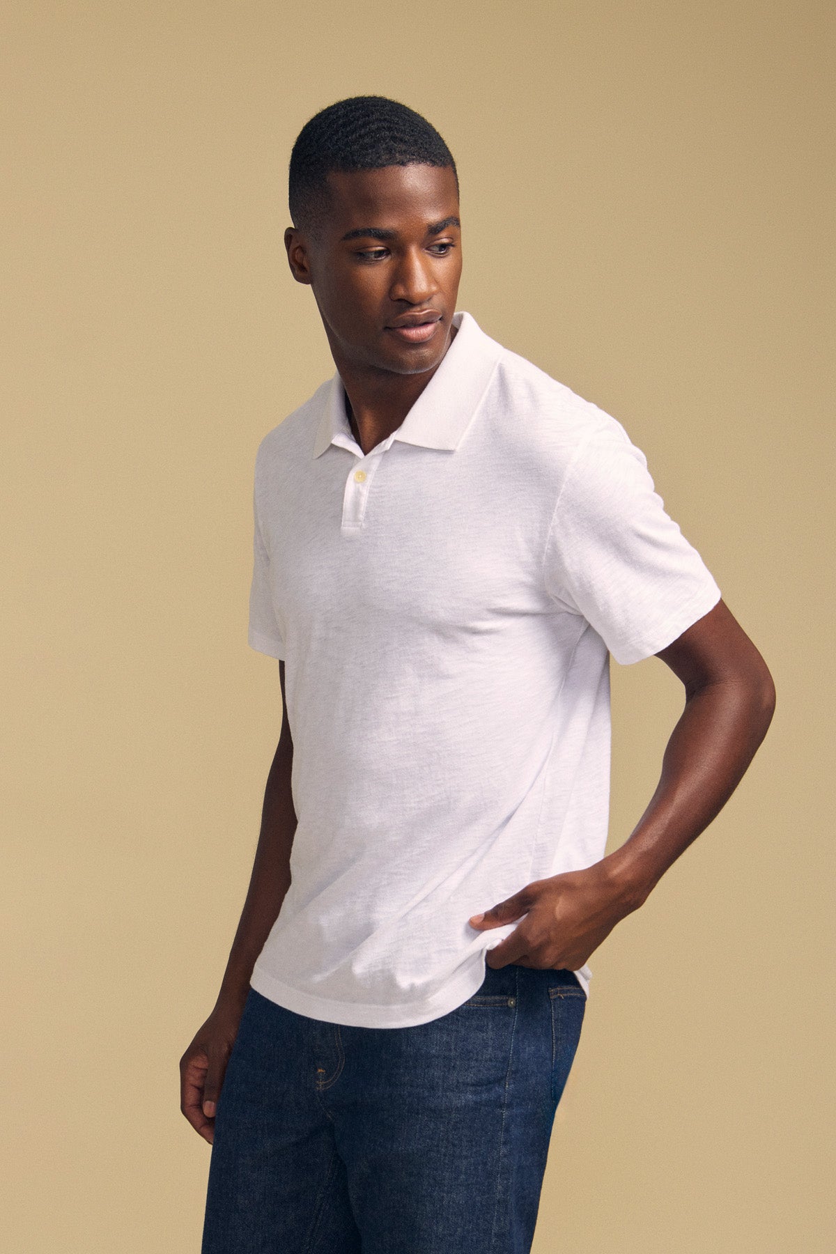 A man wearing the MORRIS POLO by Velvet by Graham & Spencer, a white short-sleeve cotton slub polo with a ribbed collar, paired with dark jeans, stands against a beige background and looks to his left.-41702098567361